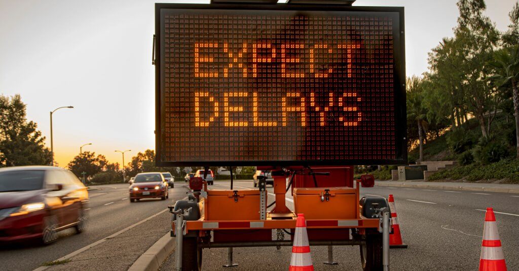 Mobile Electronic Traffic Sign stating “expect Delays” taken at sunset with traffic blurred driving past the sign and traffic cones